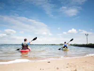 Two people paddle kayaks on Lake Couchiching from a sandy shoreline in Orillia, with calm water, blue skies, and a distant waterfront in view.