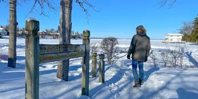 A person walks along a snowy waterfront path in Orillia beside a wooden sign, with Lake Couchiching and winter scenery in the background.