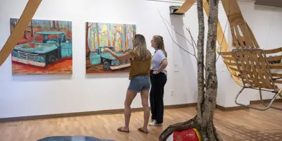 Two women view colourful paintings in an Orillia art gallery, standing beside a sculptural installation with birch trees and suspended wooden elements.