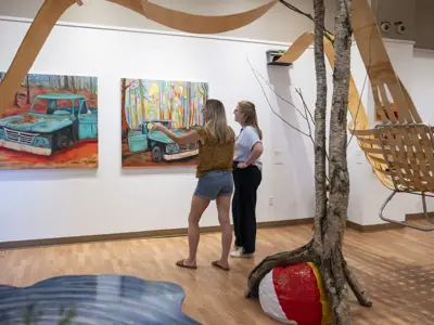Two women view colourful paintings in an Orillia art gallery, standing beside a sculptural installation with birch trees and suspended wooden elements.