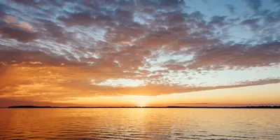 The sun sets over Lake Couchiching, casting warm orange light across calm water while scattered clouds glow in shades of gold, pink, and blue along the horizon.