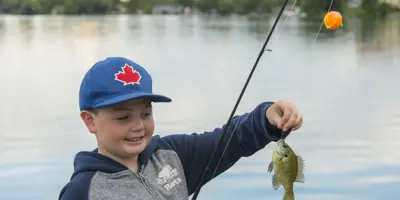 Young child smiling while holding a small fish on a fishing line by the lake, wearing a blue hat with a red maple leaf and standing near calm water
