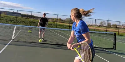 Two people playing tennis on an outdoor court, with one player preparing to hit the ball over the net on a sunny day