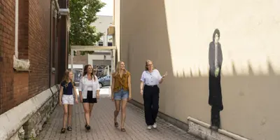 Three women and a young girl walking through an alleyway looking at a public art painting of Arthur Shilling