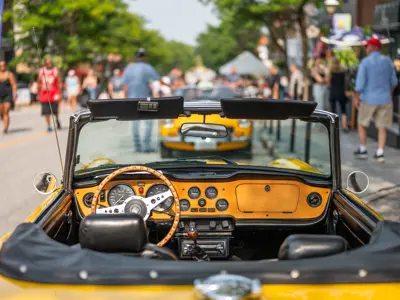 View from inside a yellow vintage convertible parked on a lively downtown street during a classic car show, with people walking among shops and displays in the background.