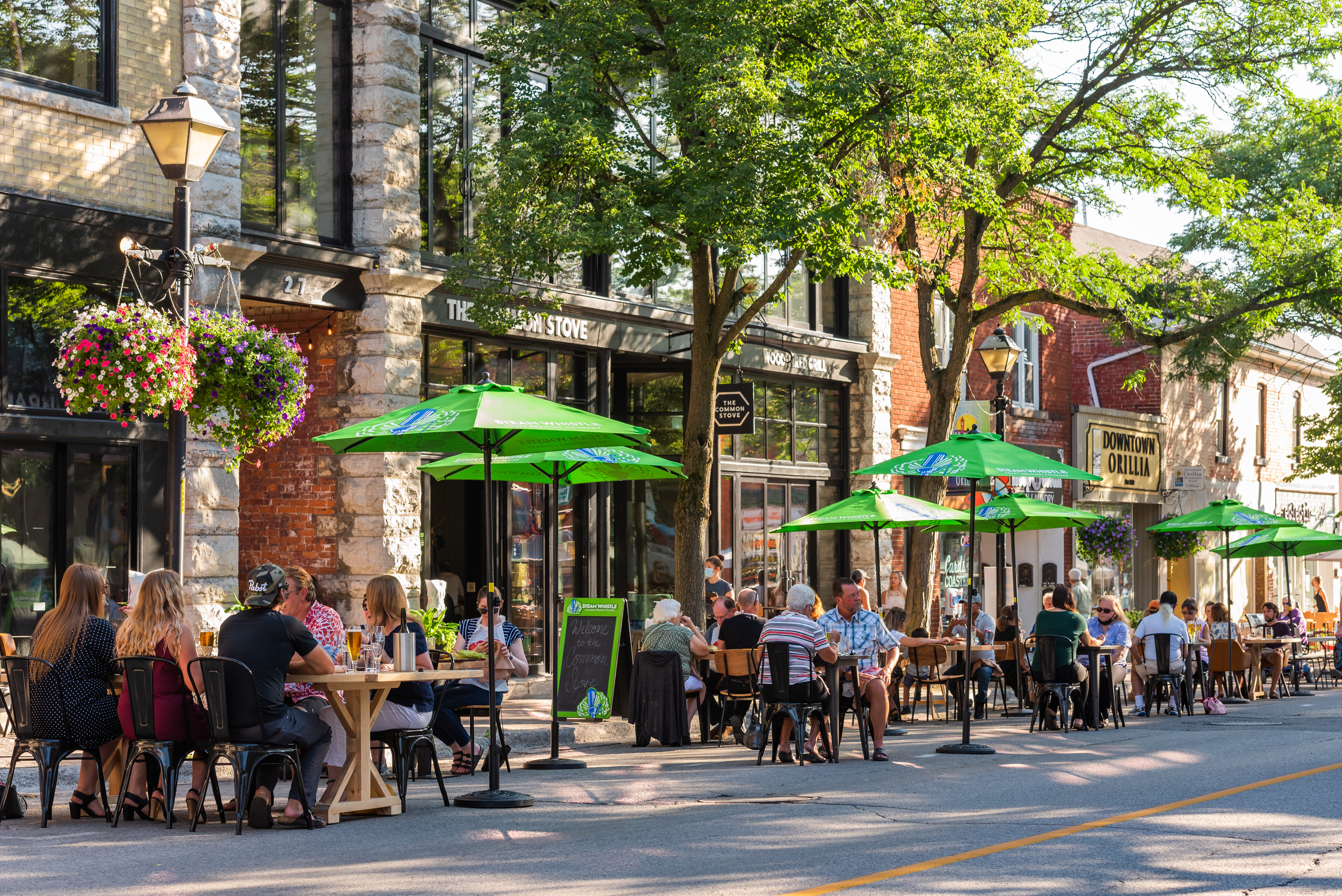 People enjoy outdoor patio dining along a lively downtown Orillia street, with green umbrellas, historic buildings, and a vibrant summer atmosphere.