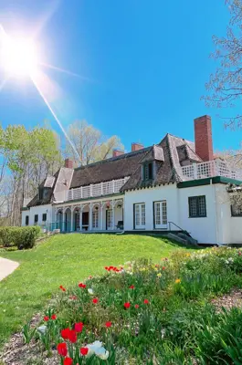 The historic Stephen Leacock Museum sits on a sunny spring day, surrounded by green lawns and blooming tulips, with tall trees and a bright blue sky overhead.