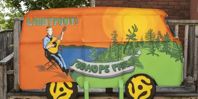 A hand painted sculpture of a vintage van depicts musician Gordon Lightfoot playing guitar by a lake