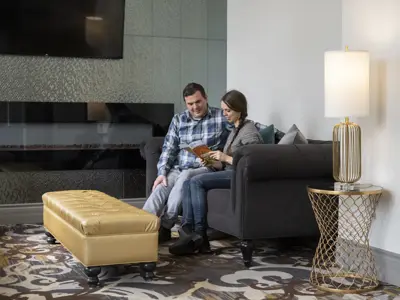A couple sits together on a comfortable couch in a modern hotel lounge, reading a brochure beside a fireplace and floor lamp in a cozy indoor setting.