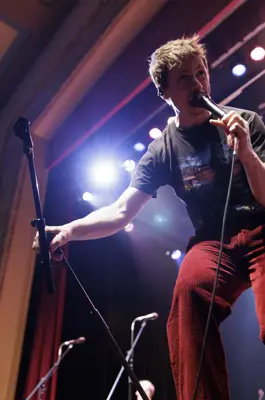 A musician sings into a handheld microphone on stage under bright concert lights, leaning toward the audience while performing live at a theatre.