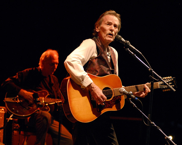 A musician sings and plays acoustic guitar on stage during a live performance in Orillia, accompanied by another guitarist under warm stage lighting.