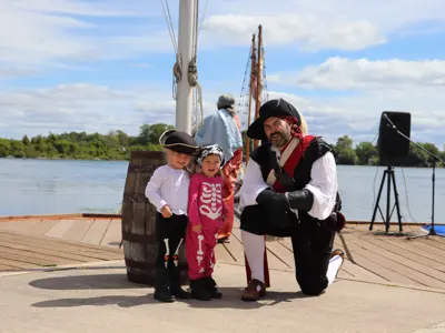 Two children dressed in skeleton costumes and pirate hats stand beside a pirate reenactor in costume on the Orillia waterfront, with a tall ship and Lake Couchiching behind them.