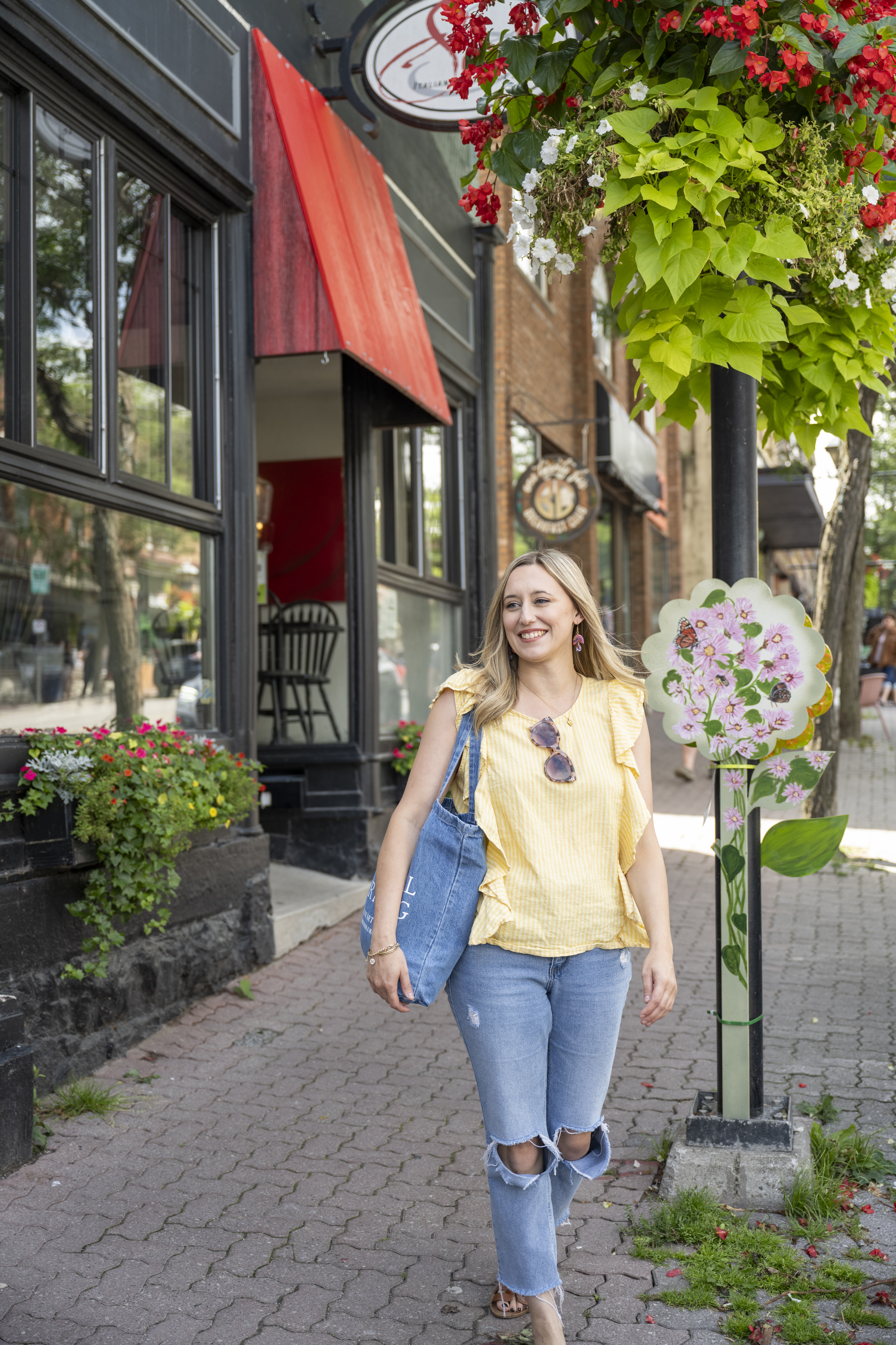 A smiling woman walks along a charming downtown sidewalk lined with storefronts, red awnings, and colourful hanging flower baskets, carrying a tote bag as she passes a decorative floral street sign.