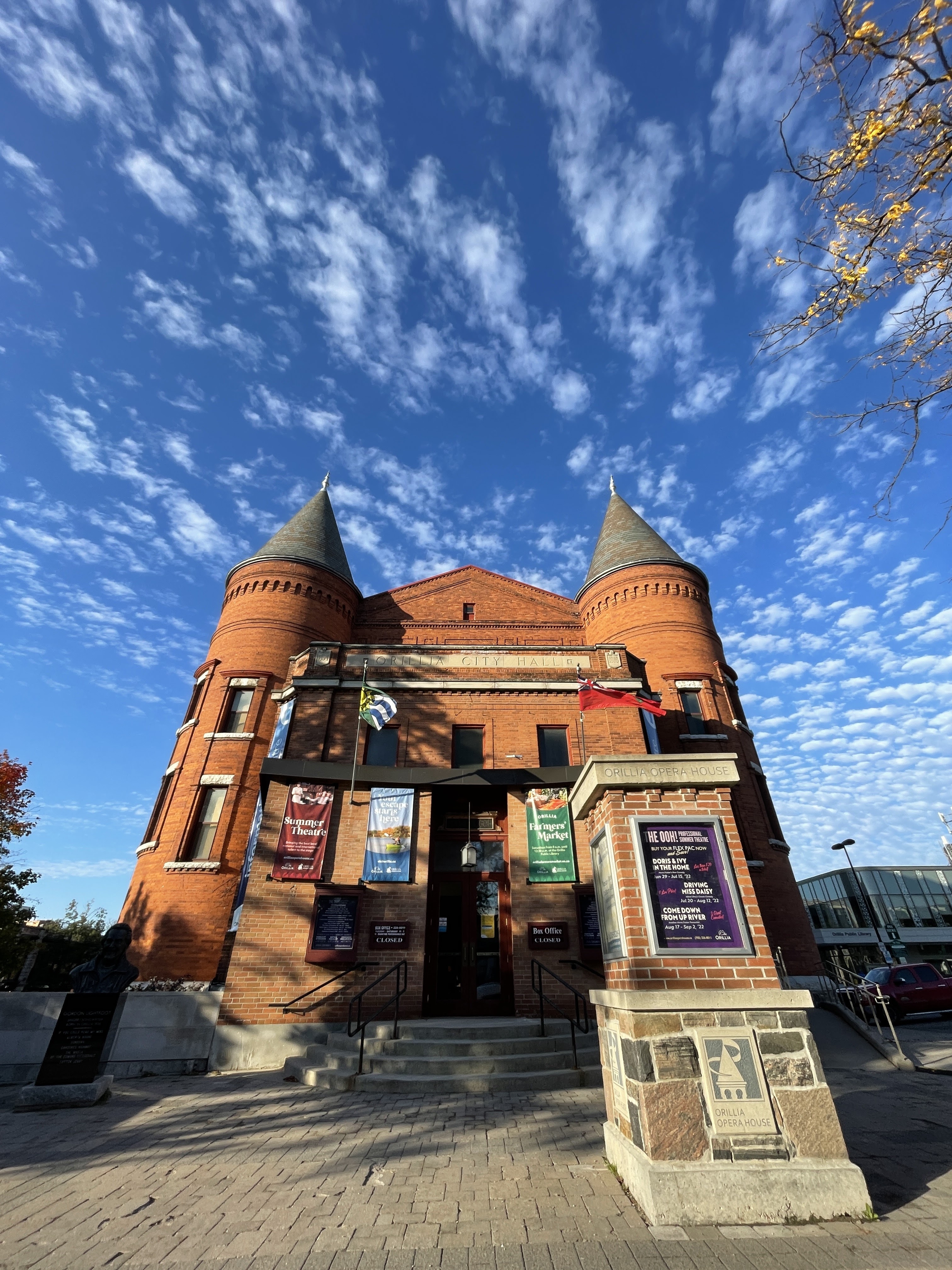 Front view of the Orillia Opera House with its red brick façade, twin turrets, and theatre banners beneath a bright blue sky with scattered clouds.