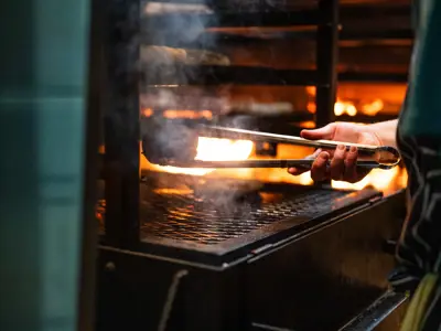 Close-up of a hand using tongs to place food on a grill over an open flame, with smoke rising in a warm, restaurant kitchen setting.