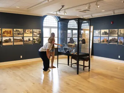 Two women lean in to examine artifacts inside a glass display case at an Orillia museum, surrounded by historic photographs on dark gallery walls.