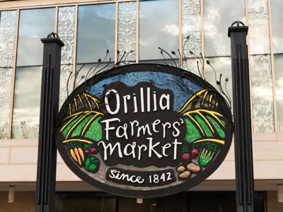 Colourful sign for the Orillia Farmers’ Market reading “Since 1842,” featuring illustrations of produce and local goods in front of a building façade.