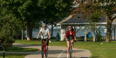 Two cyclists ride along a paved path in an Orillia waterfront park, with green trees, open space, and a gazebo in the background.