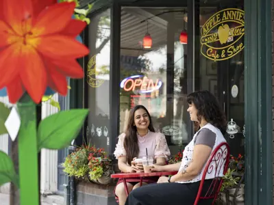Two women sit at a small red patio table outside Apple Annie’s Café & Shop, smiling and chatting with iced drinks while a bright red flower is blurred in the foreground and an open sign glows in the window.