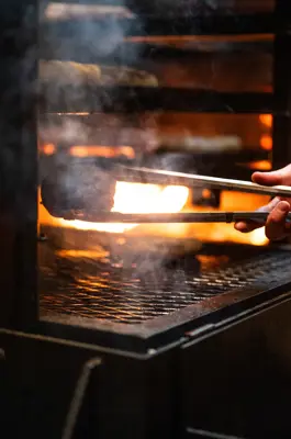 A cook uses metal tongs to turn food on a hot grill as flames and smoke rise from an open-flame barbecue in a restaurant kitchen.