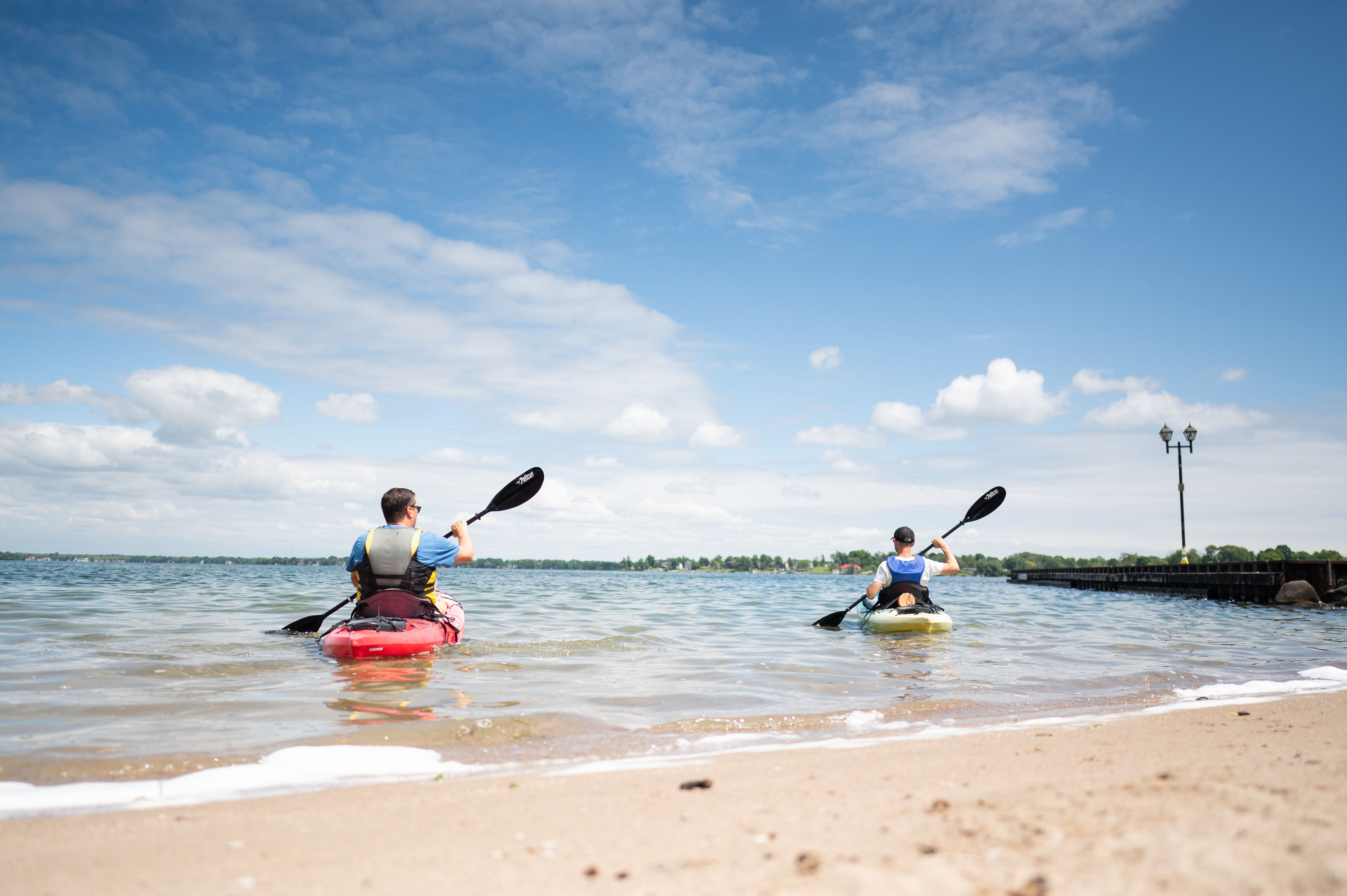 Two people launch kayaks from a sandy beach into Lake Couchiching in Orillia, paddling into calm water under a bright blue sky.