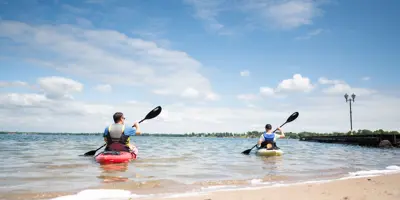 Two people launch kayaks from a sandy beach into Lake Couchiching in Orillia, paddling into calm water under a bright blue sky.